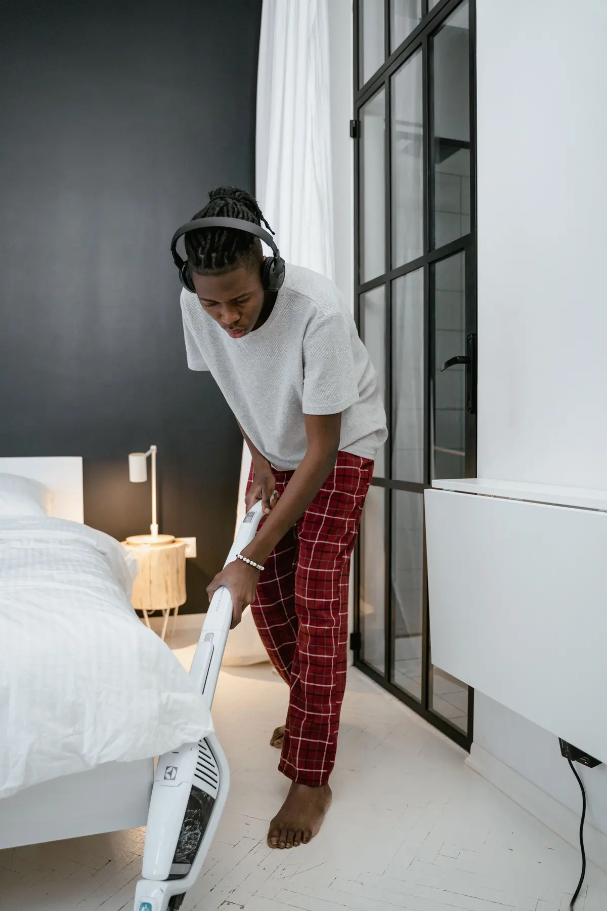 Barefoot man in headphones, a gray T-shirt and red plaid pajama pants vacuuming a white plank floor with a cordless stick vacuum beside a white bed, with a black-framed glass door behind him